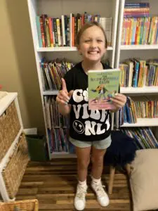 A young girl holding up a book in front of a bookshelf.